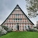 An A shaped house with an edifice featuring wood that separates squares of patterned bricks, an open front door, blooming flowers and a manicured lawn
